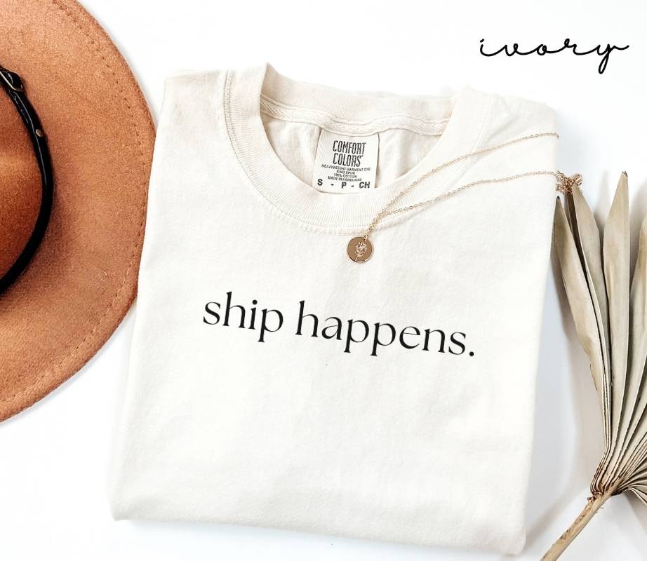 White (ivory) t-shirt with 'ship happens.' text, brown hat, and dried leaves on a white background