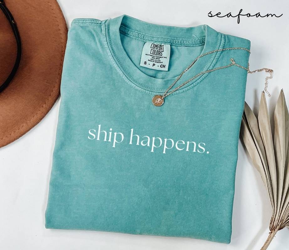 Folded teal (seafoam) t-shirt with 'ship happens.' text, surrounded by a hat and dried leaves on a white background.