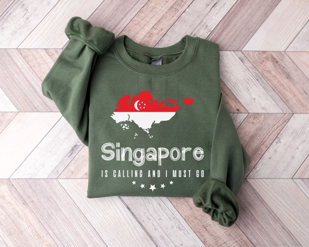 A millitray green sweatshirt featuring a design inspired by Singapore is laid out on a wooden floor.
