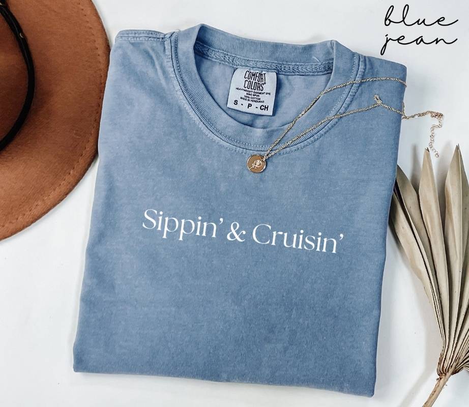 Folded blue jean t-shirt with 'Sippin' & Cruisin'' text, next to a hat and dried leaves on a white background.
