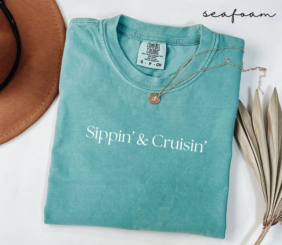 Folded teal (seafoam) t-shirt with 'Sippin' & Cruisin'' text, surrounded by a hat and dried leaves on a white background.