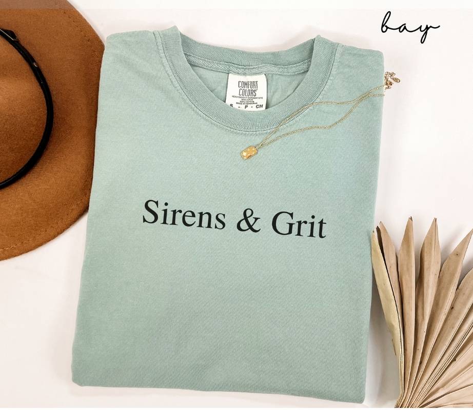 Folded light green (bay) t-shirt with 'Sirens & Grit' text, next to a brown hat and dried leaves on a white background.