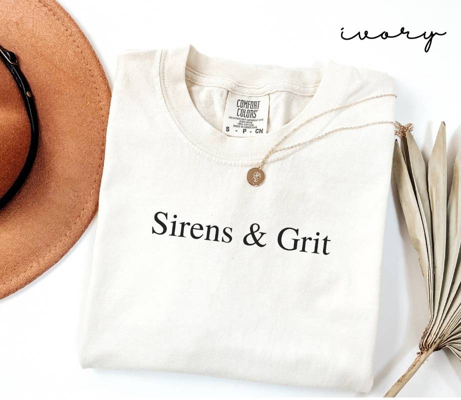 White (ivory) t-shirt with 'Sirens & Grit' text, accompanied by a hat and dried plant on a white background.