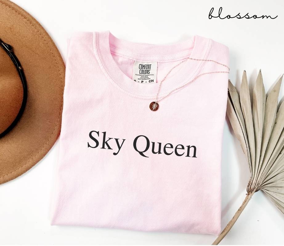 Pink (blossom) t-shirt with 'Sky Queen' text, brown hat, and dried plant on a white background