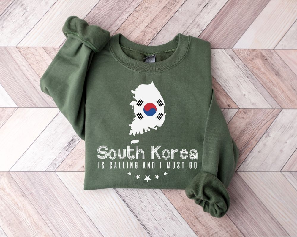 A millitray green sweatshirt featuring a design inspired by South Korea is displayed on a wooden floor.