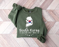 A millitray green sweatshirt featuring a design inspired by South Korea is displayed on a wooden floor.