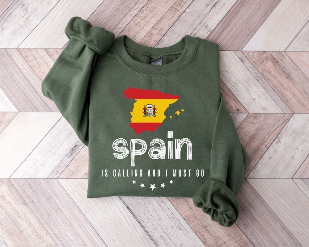 A military green sweatshirt featuring the Spanish flag and text, laid out on a wooden floor.