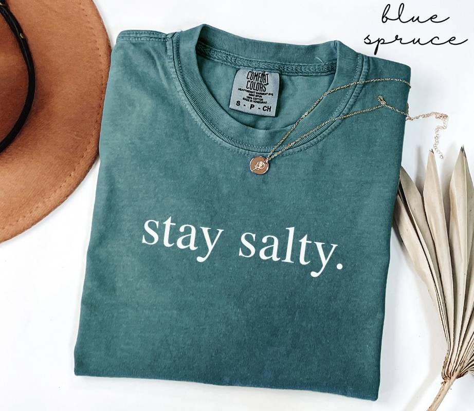 Folded teal (blue spruce) t-shirt with 'stay salty.' text, brown hat, and dried leaves on a white background