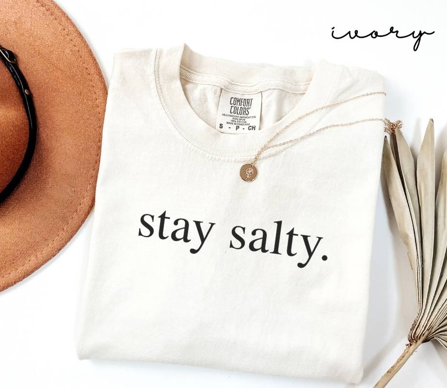White (ivory) t-shirt with 'stay salty.' text, placed on a white surface with a hat and dried plant.