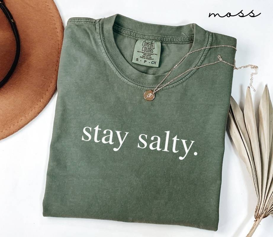 Folded green (moss) t-shirt with 'stay salty.' text, next to a brown hat and dried leaves on a white background.