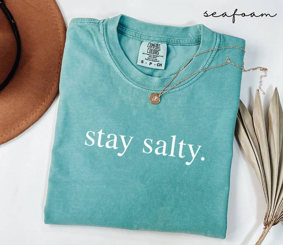 Folded teal (seafoam) t-shirt with 'stay salty.' text, surrounded by a hat and dried leaves on a white background.