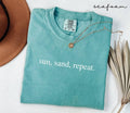Folded teal (seafoam) t-shirt with 'sun, sand, repeat.' text, surrounded by a hat and dried leaves on a white background.