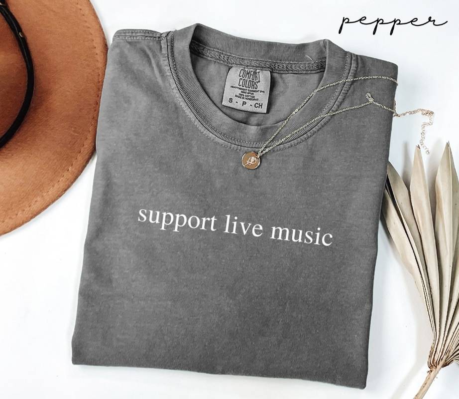 Folded gray (pepper) t-shirt with 'support live music' text, placed on a white surface with a hat and dried leaves.