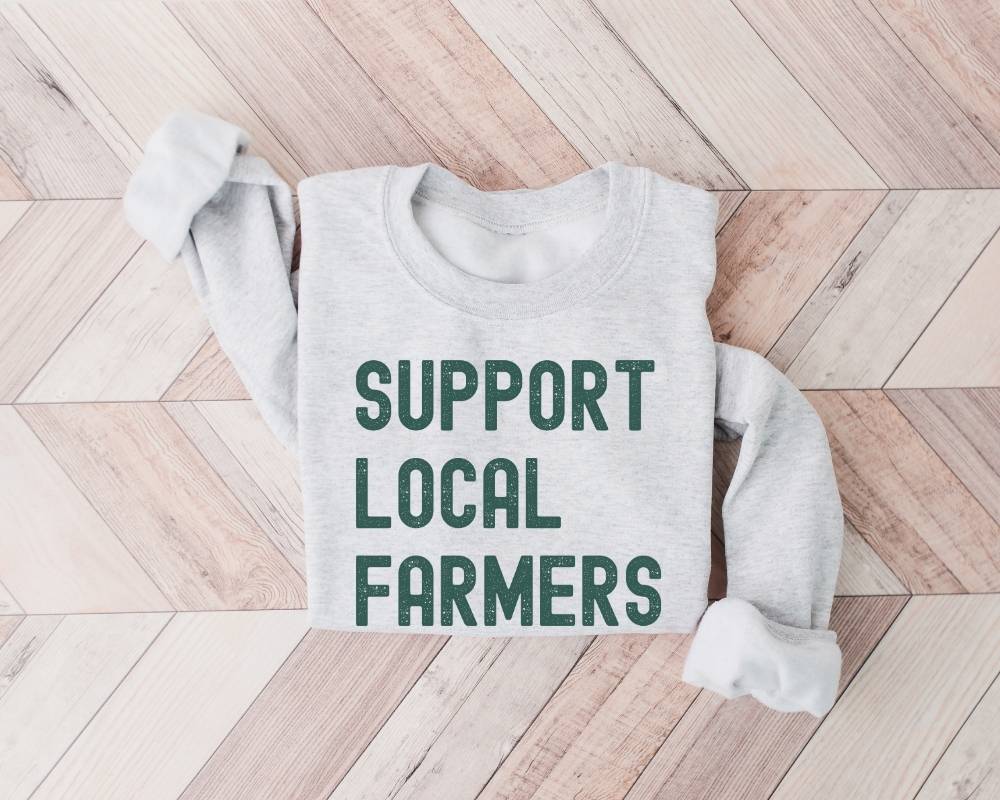 Ash (Gray) sweatshirt with 'Support Local Farmers' retro dark green text on a wooden floor