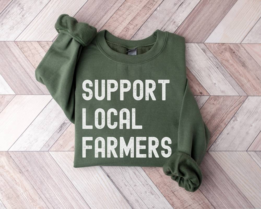 Military Green sweatshirt with 'Support Local Farmers' retro text on a wooden floor.