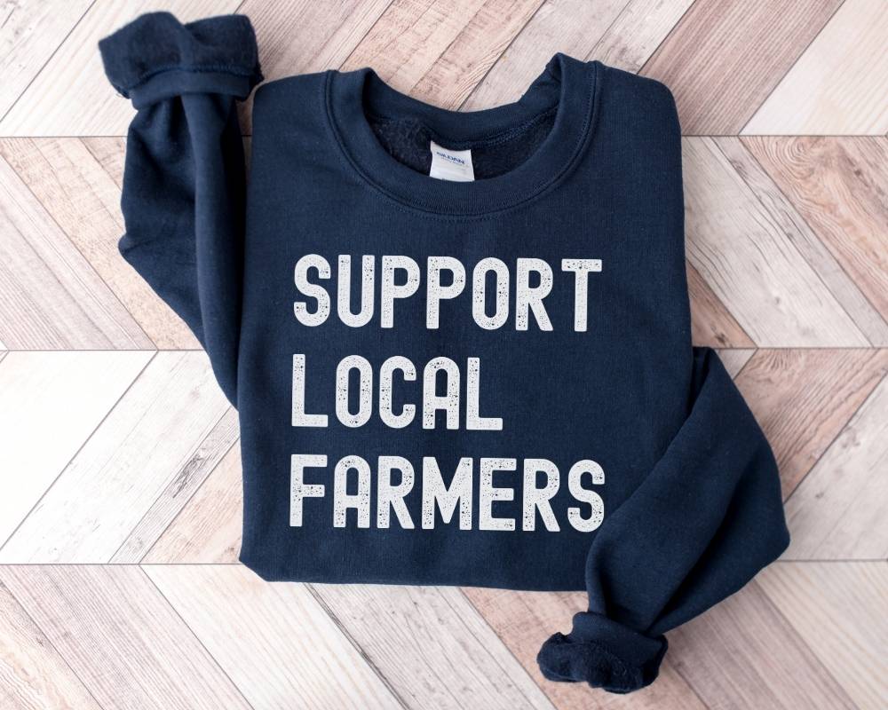 Navy blue sweatshirt with 'Support Local Farmers' text on a wooden floor.