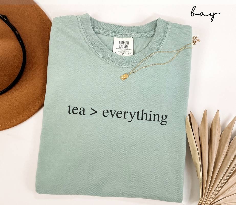Folded light green (bay) t-shirt with 'tea > everything' text, next to a brown hat and dried leaves on a white background.