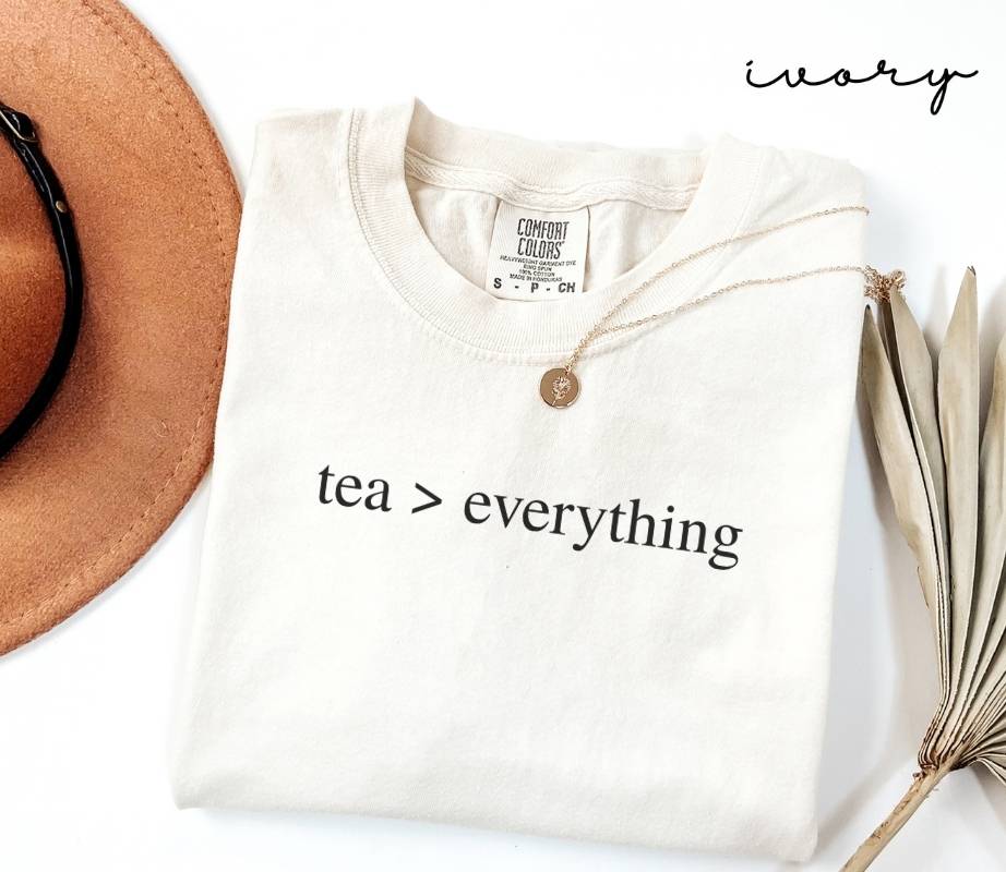 White (ivory) t-shirt with 'tea > everything' text, next to a brown hat and dried leaves on a white background.