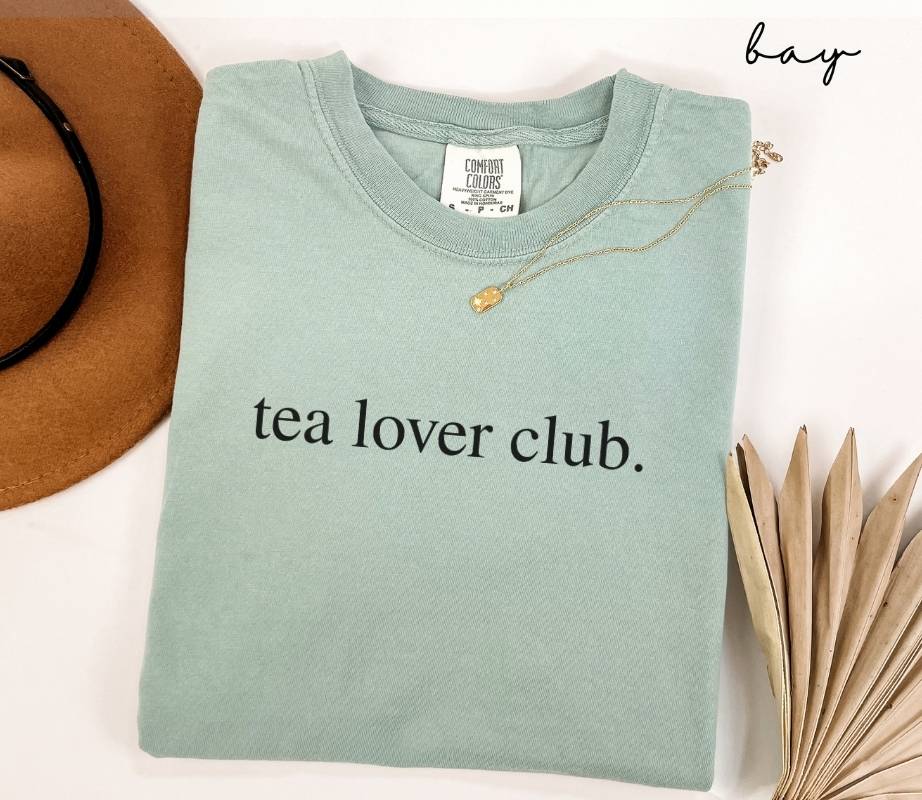 Light green (bay) t-shirt with 'tea lover club' text, placed on a white surface with a hat and dried leaves.