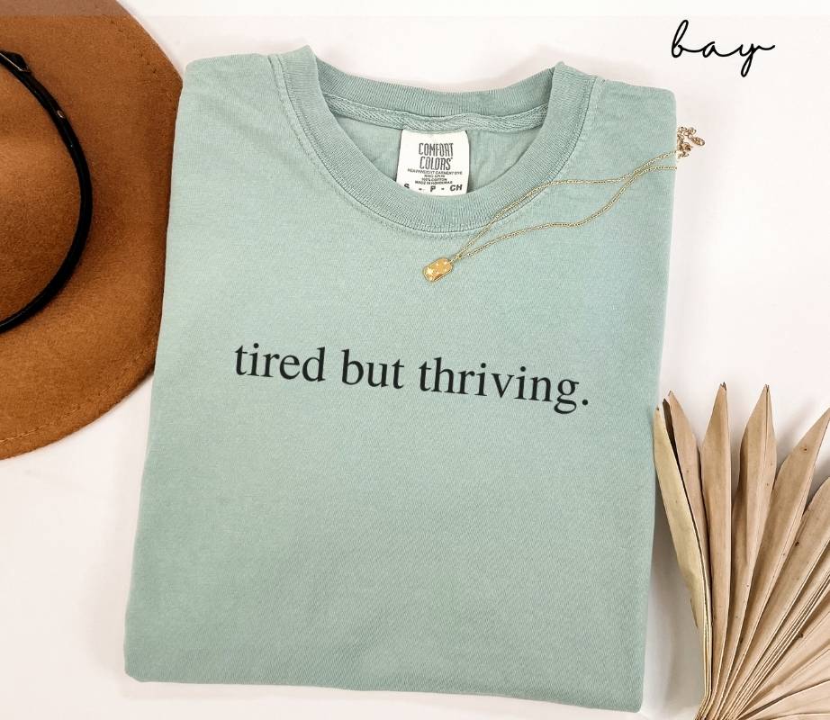 A folded light green t-shirt with the text 'tired but thriving,' placed next to a brown hat and dried leaves on a white background.