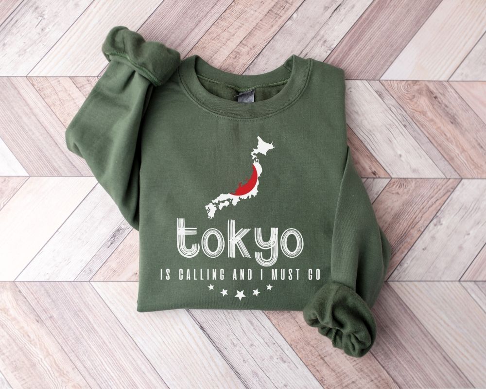 Military green sweatshirt featuring 'Tokyo is Calling and I Must Go' text along with a map design on a wooden floor.