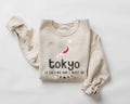 A beige (sand) sweatshirt featuring the phrase "Tokyo is Calling" along with a graphic on a white background.