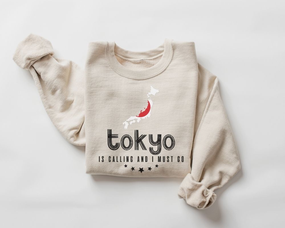 A beige (sand) sweatshirt featuring the phrase "Tokyo is Calling" along with a graphic on a white background.