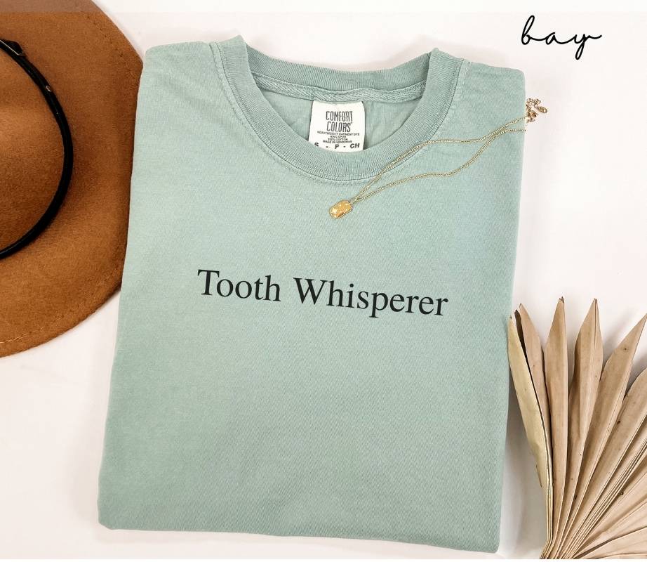 Folded light green (bay) t-shirt with 'Tooth Whisperer' text, next to a brown hat and dried leaves on a white background.