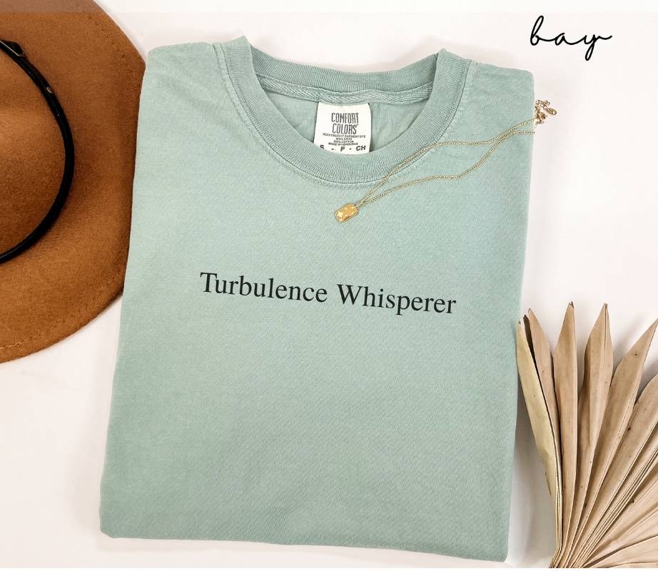 Folded light green (bay) t-shirt with 'Turbulence Whisperer' text, next to a brown hat and dried leaves on a white background.