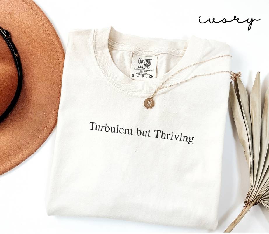 White (ivory) t-shirt with 'Turbulent but Thriving' text, accompanied by a hat and dried plant on a white background.