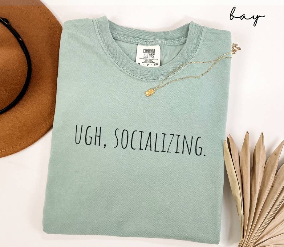 Light green t-shirt with the text 'UGH, SOCIALIZING.' displayed on a white surface alongside a hat and dried leaves.