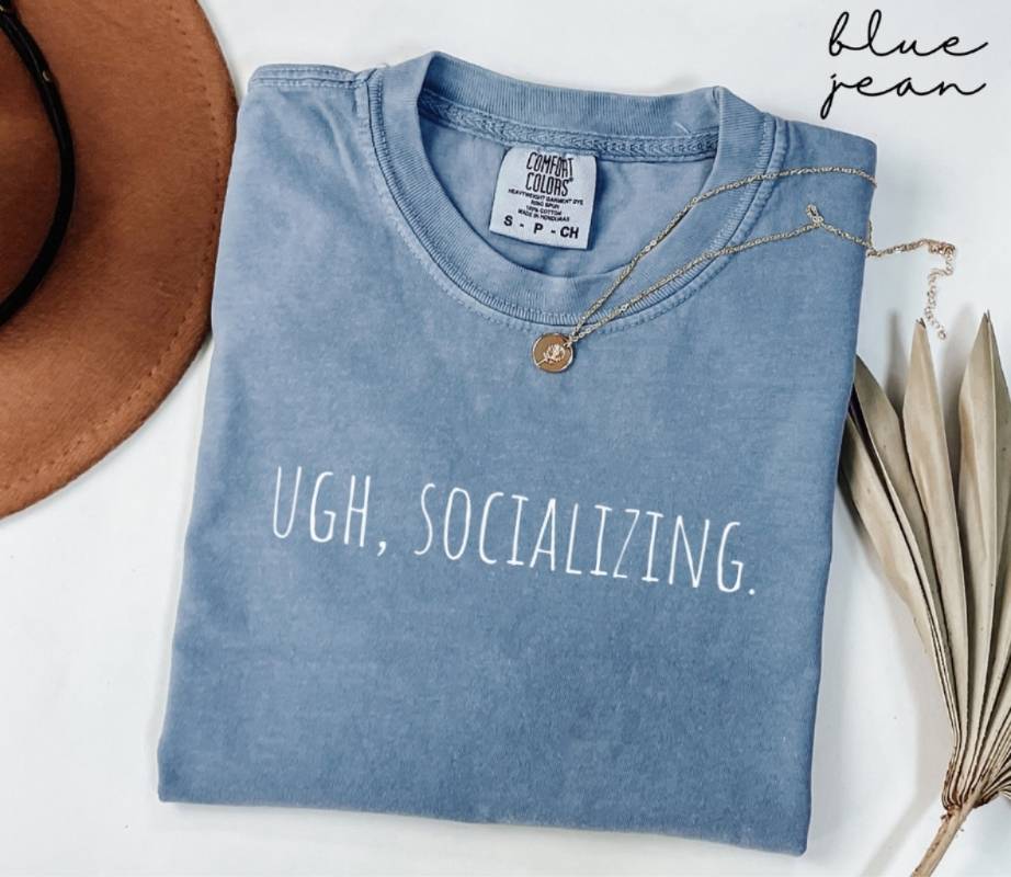 A folded blue t-shirt displaying the phrase 'UGH, SOCIALIZING,' accompanied by a hat and a plant, placed on a white background.