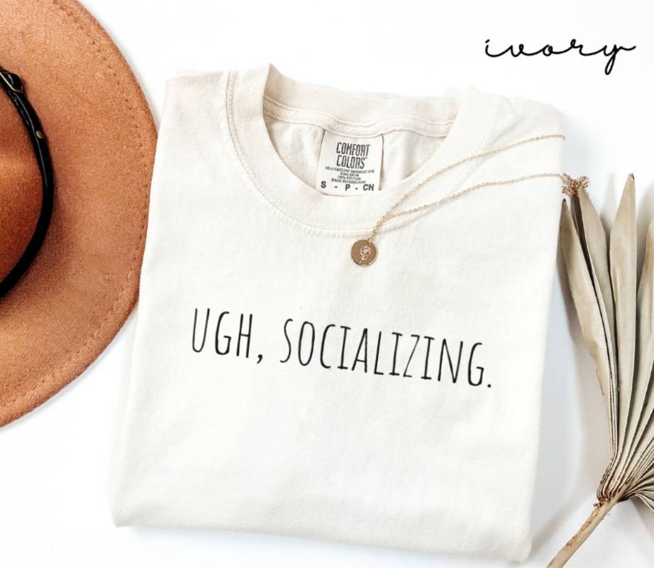 White t-shirt with the text 'UGH, SOCIALIZING.', a brown hat, and a dried plant on a white background.