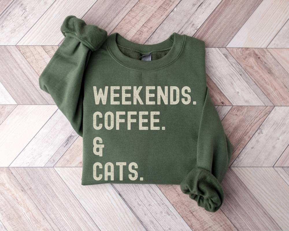 Military green sweatshirt with 'Weekends. Coffee. & Cats.' text on a wooden floor.
