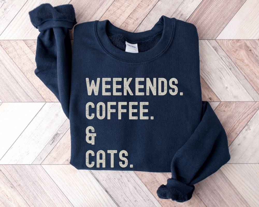 Navy blue sweatshirt with 'Weekends. Coffee. & Cats.' text on a wooden floor.
