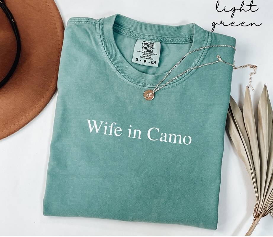 Folded light green t-shirt with 'Wife in Camo' text on a white surface with a hat and dried leaves.