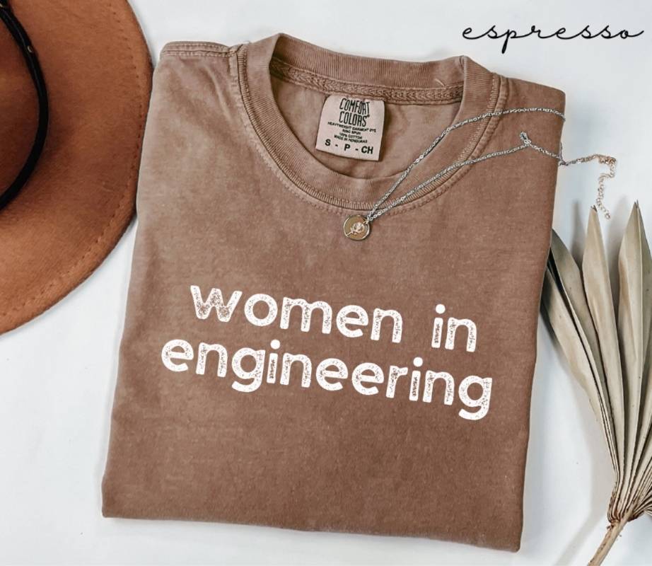 Folded brown (espresso) t-shirt with 'women in engineering' text on a white surface with a hat and dried plant.