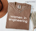 Folded brown (espresso) t-shirt with 'women in engineering' text on a white surface with a hat and dried plant.