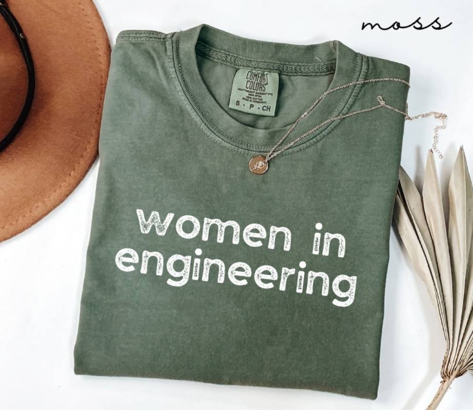 Folded green (moss) t-shirt with 'women in engineering' text on a white surface with a hat and dried leaves.