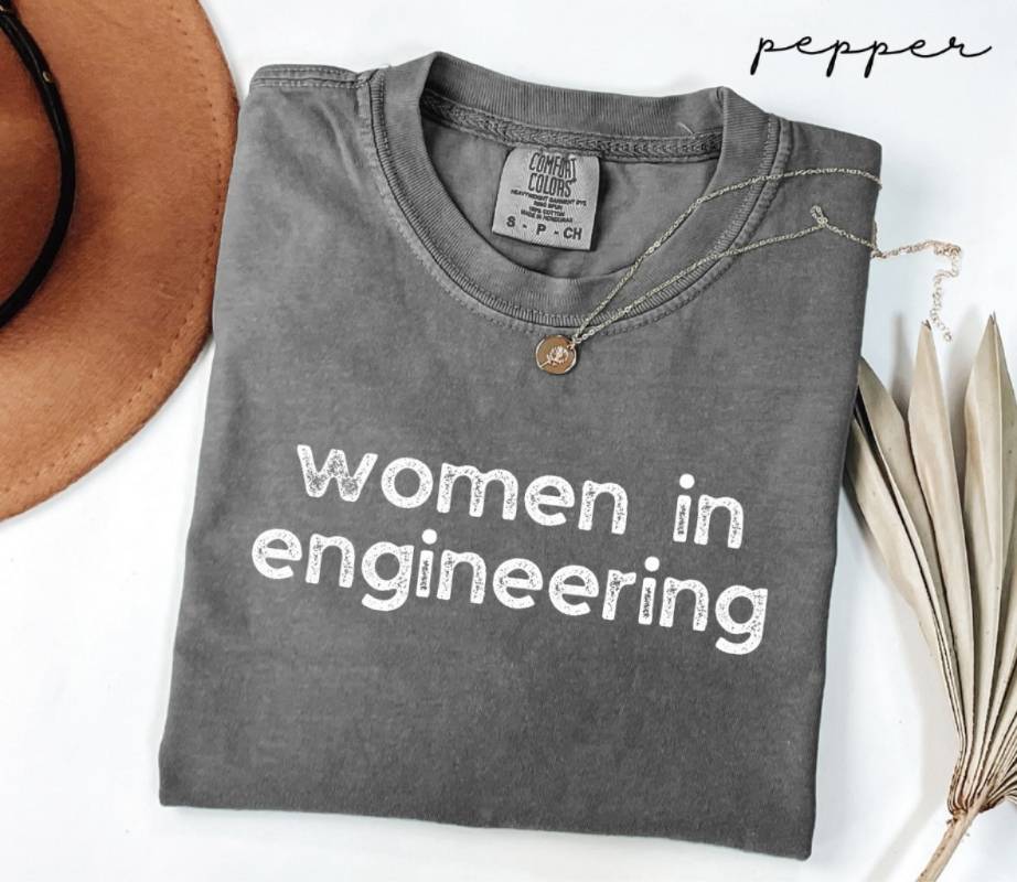 Gray (pepper) t-shirt with 'women in engineering' text on a white surface with a hat and dried leaves.