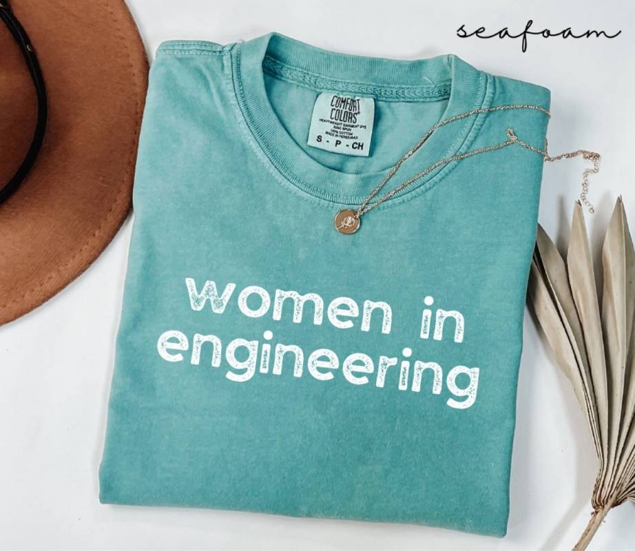 Folded teal (seafoam) t-shirt with 'women in engineering' text on a white surface with a hat and dried plant.