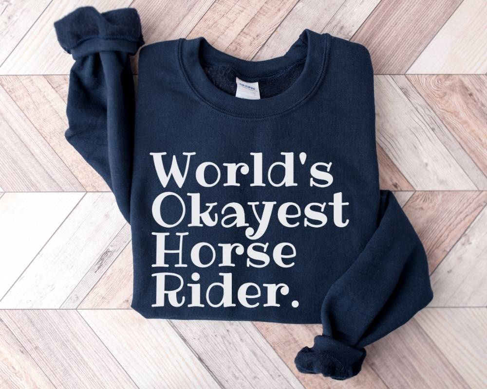 Navy blue sweatshirt with 'World's Okayest Horse Rider' text on a wooden floor.